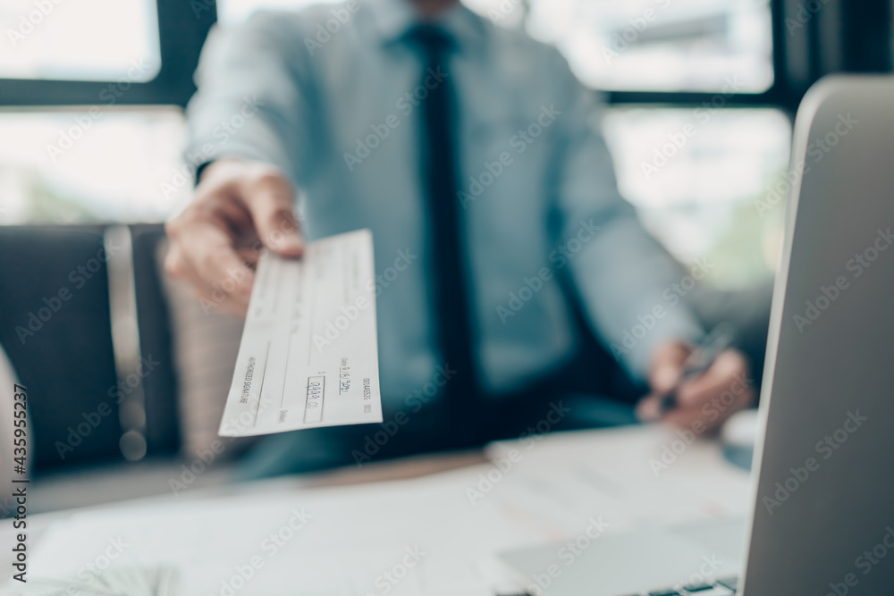 A Businessperson's hand giving cheque to customer and dollar bill, coin, laptop and graph chart on the desk at office. Payment by check, paycheck, payroll concept.