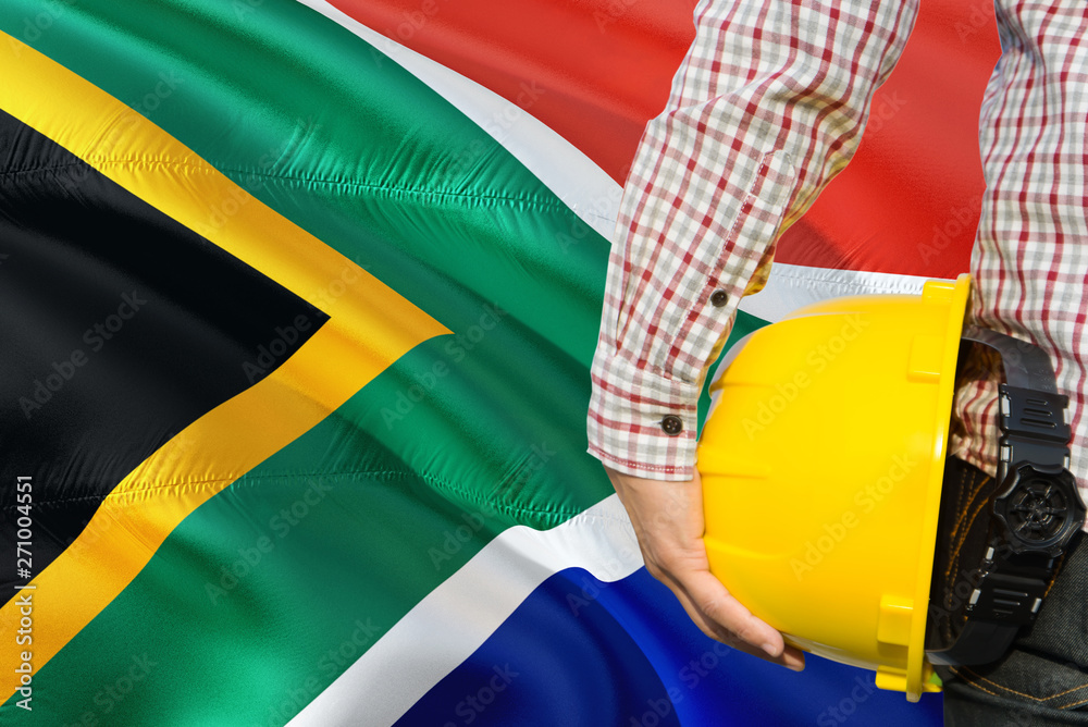 Engineer is holding yellow safety helmet with waving South Africa flag background. Construction and building concept.