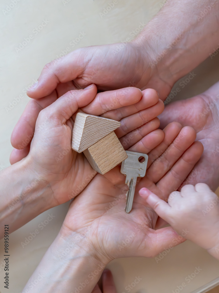 A child hand points to a front door key next to a small wooden toy house in the open hands of his parents.