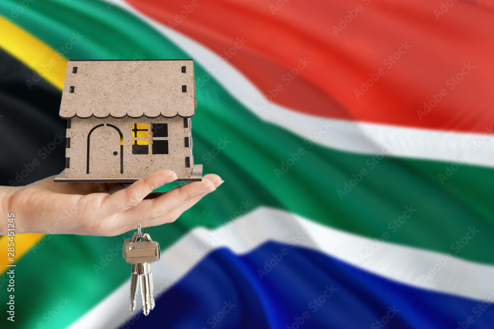 Woman hand holding wooden house and keys with hand. South Africa flag with concept of rent, purchase, insurance, building real estate, eco house.