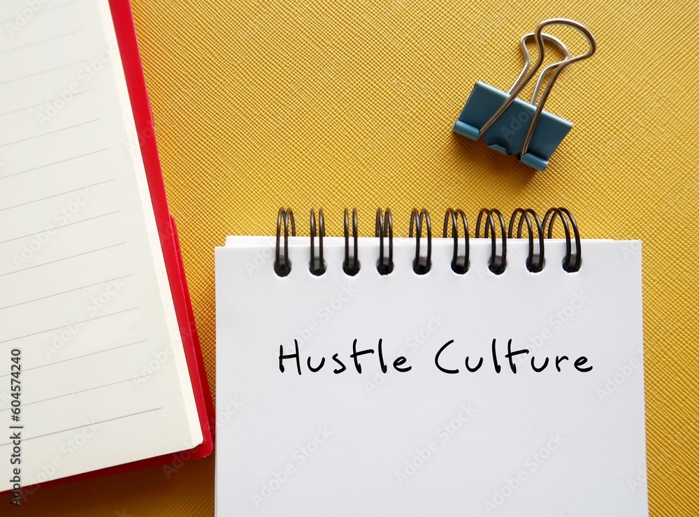 Notbook on yellow background with handwritten text HUSTLE CULTURE - lifestyle where career has become first priority, state of constant overworking to the point where it becomes work life imbalance